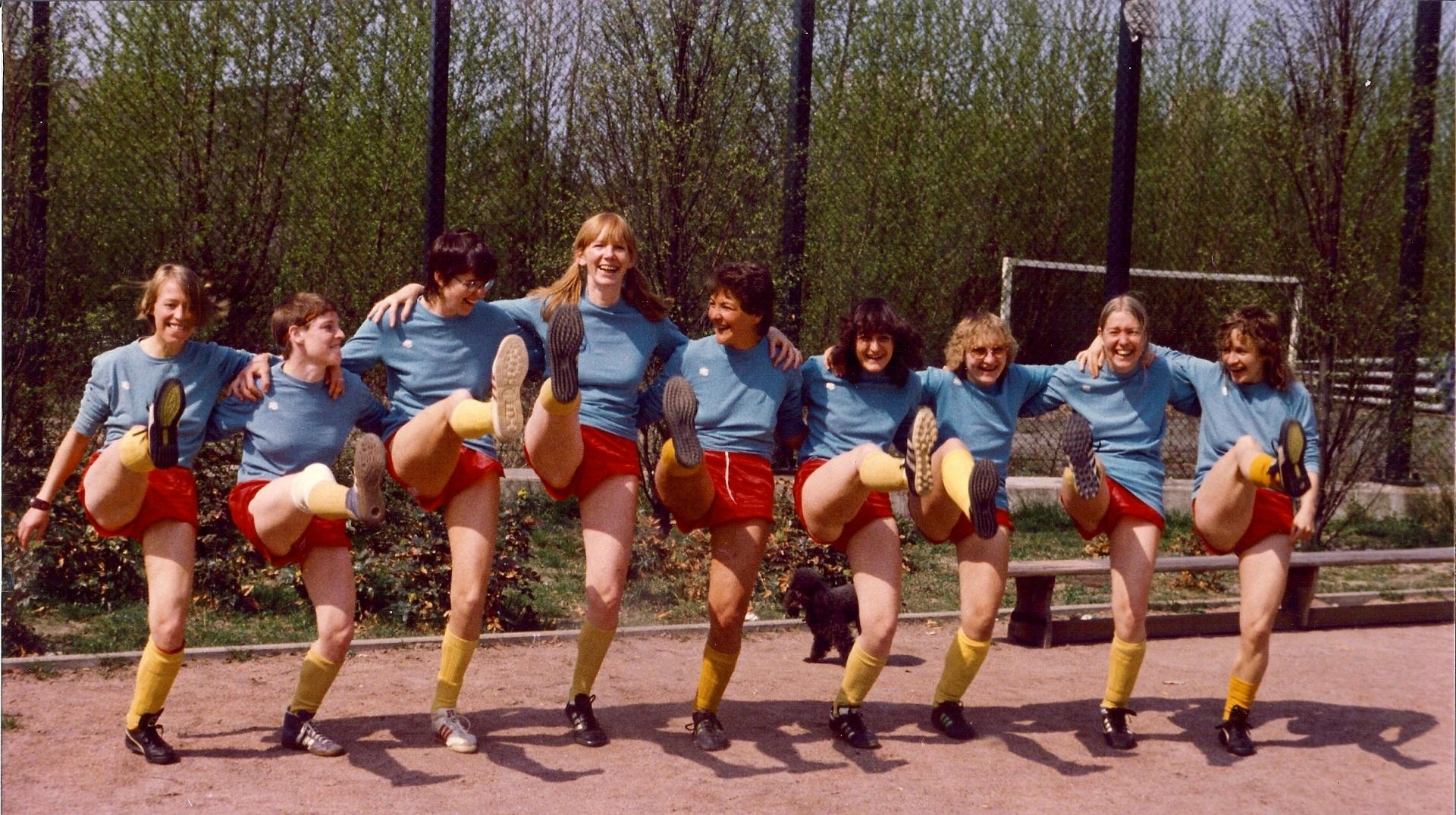 Neun junge Frauen stehen Arm in Arm auf einem Sportplatz und schwingen gleichzeitig das rechte Bein hoch.