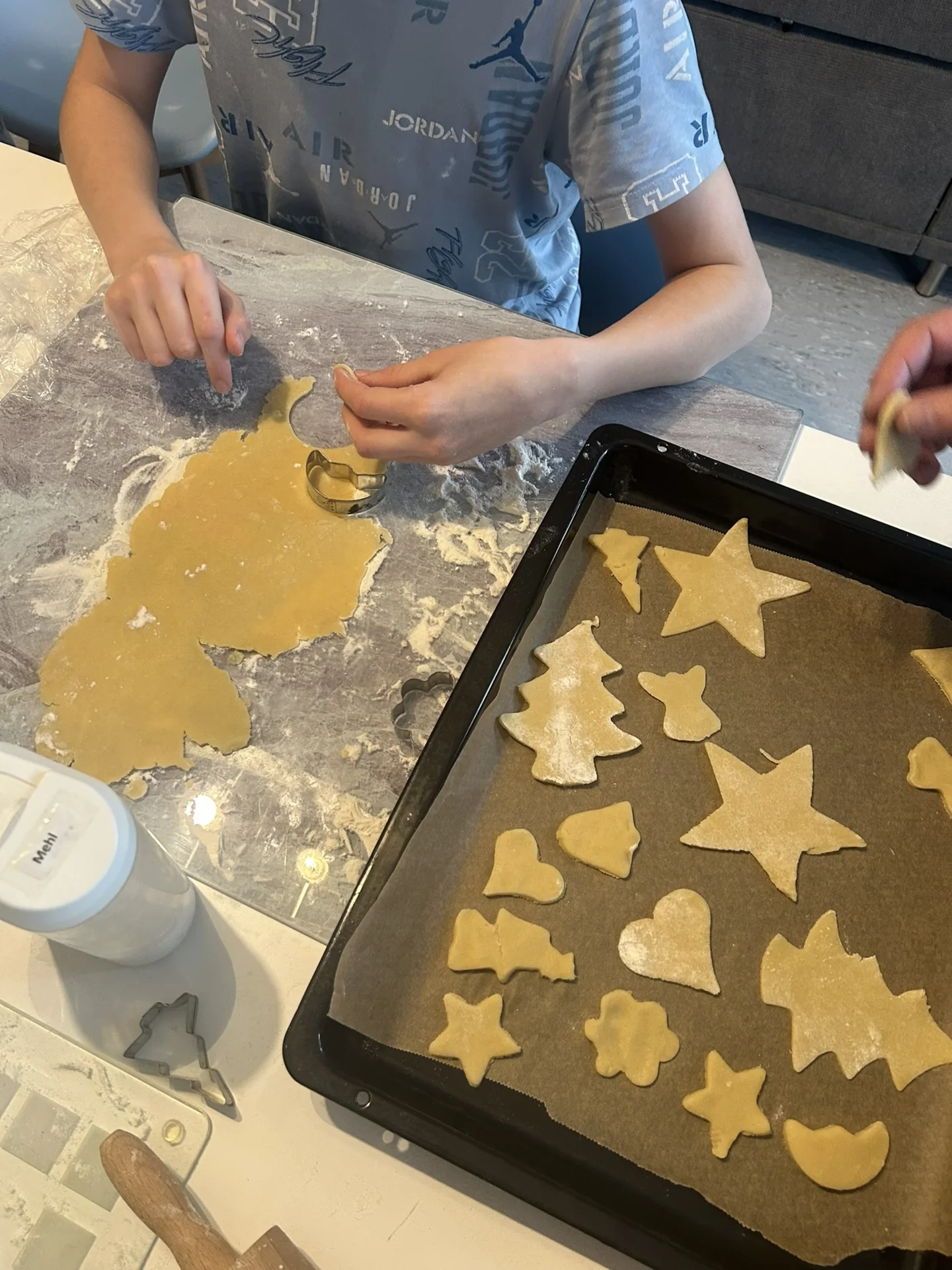 fertig ausgestochene Plätzchen liegen auf einem Blech, daneben sticht ein Junge noch weitere aus Plätzchenteig aus
