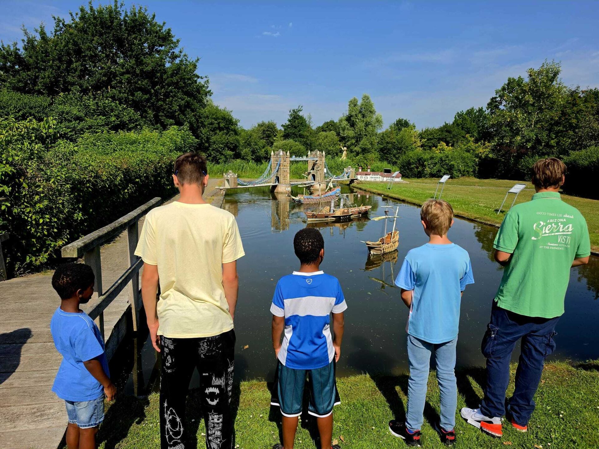fünf Kinder stehen vor einem See, in welchem sich kleine Schiffe und ein kleiner Nachbau der Towerbridge befinden, mit dem Rücken zum Betrachter