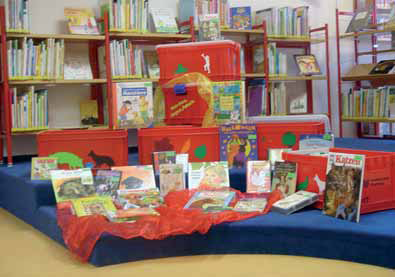Auf dem Bild sieht man vier rote Boxen, die teilweise mit verschiedenen Motiven beklebt sind, und davor stehen bzw. liegen einige Bücher.