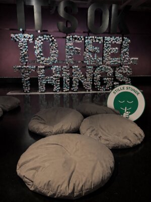 Das Bild zeigt einen ruhigen und abgedunkelten Ausstellungsbereich mit mehreren großen Sitzkissen auf dem Boden. Im Hintergrund steht der Schriftzug "It´s OK to feel things", auf dem Klebezettel platziert sind. Rechts ist das Symbol der "Stillen Stunde" zu sehen. Die Atmosphäre lädt Besucher:innen zum Ausruhen und Entspannen ein. (Bild vergr&ouml;&szlig;ern)