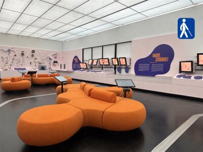 Das Bild zeigt einen modernen Ausstellungsraum mit viel Platz und einer ruhigen Atmosphäre. Im Vordergrund stehen mehrere große, orangefarbene Sitzinseln mit weichem, runden Formen, während sich an den Wänden interaktive Stationen, Bildschirme und Informationstafeln befinden. Rechts oben im Bild ist gut sichtbar ein blaues Piktogramm mit einer Person und einem Langstock angebracht, das auf das Angebot für blinde und seheingeschränkte Besucher:innen hinweist. Der Raum wirkt hell und übersichtlich. Man erkennt auch Teile des Blindenleitsystems auf dem Fußboden. (Bild vergr&ouml;&szlig;ern)
