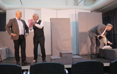 Ein Mann zeigt sehr aufgeregt, einem anderen Mann ein Papier. Der andere Mann wirkt bedrohlich. Ein anderer Mann, der hinter den beiden ist, wühlt in Kleidern rum. Im Hintergrund die Kulisse der Theaterbühne. (Bild vergr&ouml;&szlig;ern)
