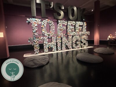 Das Bild zeigt einen ruhigen und abgedunkelten Ausstellungsbereich mit mehreren großen Sitzkissen auf dem Boden. Im Hintergrund steht der Schriftzug: It´s OK to feel things, auf dem Klebezettel platziert sind. Rechts ist das Symbol der Stillen Stunde zu sehen. Die Atmosphäre lädt Besucher:innen zum Ausruhen und Entspannen ein. (Bild vergr&ouml;&szlig;ern)